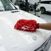 A person's hand in a red microfiber chenille wash glove covered in suds, cleaning the white hood of a car.